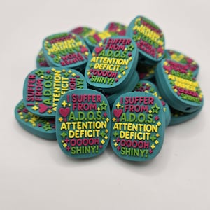 May include: A pile of teal-colored beads with the text "I SUFFER FROM A.D.O.S. ATTENTION DEFICIT OOOOH SHINY!" in yellow, pink, and green. Each bead has a heart and star design.