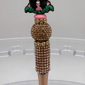 May include: A decorative pen with a rose gold barrel and a jeweled top. The pen features a charm of a woman wearing a green hat and pink dress. The pen's upper section is adorned with sparkling crystals.