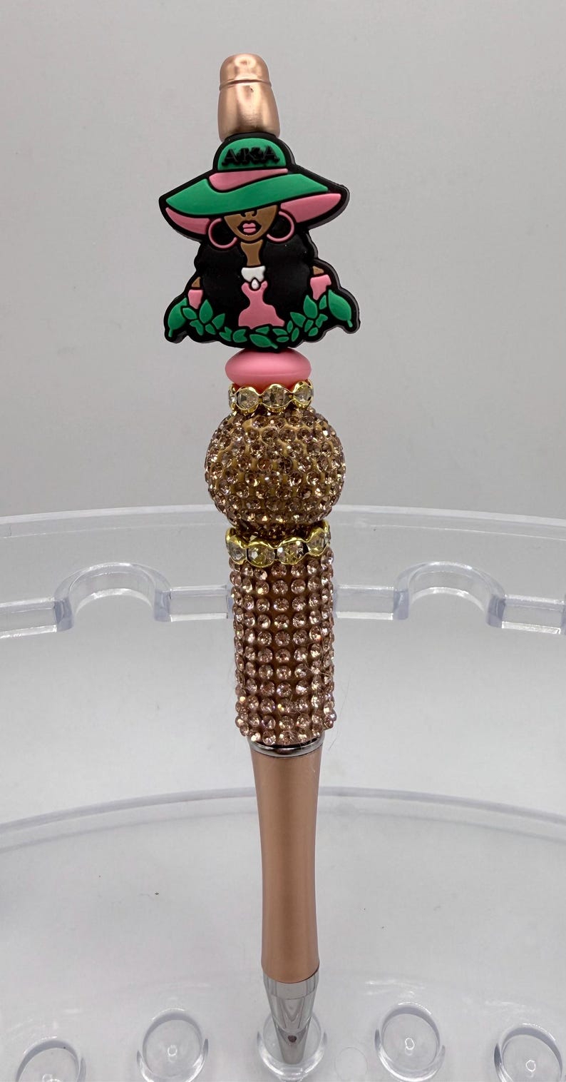May include: A decorative pen with a rose gold barrel and a jeweled top. The pen features a charm of a woman wearing a green hat and pink dress. The pen's upper section is adorned with sparkling crystals.