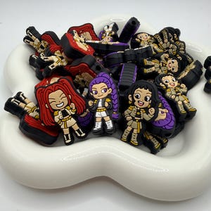 May include: A collection of colorful cartoon character shoe charms in a white, wavy dish. The charms feature various characters with red, purple, and black hair, wearing outfits in shades of tan, white, and black. The charms are made of rubber.