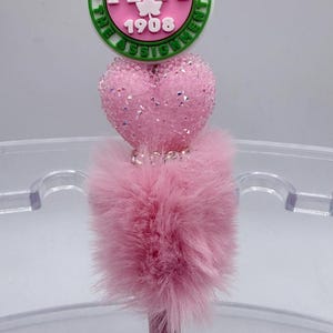 May include: Pink pen with a decorative top featuring a green and white circular emblem with the letters "AKA" and the number "1908". The pen has a pink, fluffy base and a heart-shaped embellishment.