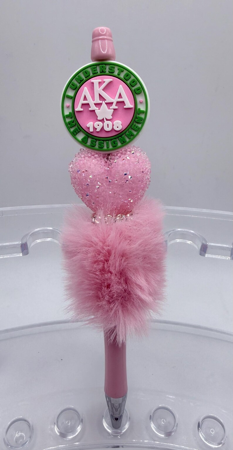 May include: Pink pen with a decorative top featuring a green and white circular emblem with the letters "AKA" and the number "1908". The pen has a pink, fluffy base and a heart-shaped embellishment.