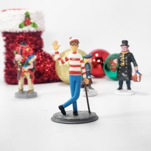 May include: A figurine of a character in a red and white striped top, blue trousers, and glasses, waving. Other figurines and Christmas baubles are in the background, including a red stocking and a gold bauble. The figurine is on a grey base.