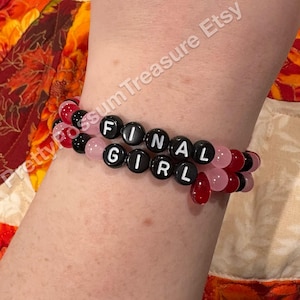 Op de afbeelding: Twee armbanden met de woorden "FINAL GIRL" gespeld in witte letters op zwarte kralen. De armbanden hebben ook roze, rode en zwarte kralen. De armbanden worden om een pols gedragen.
