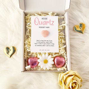 K&ouml;nnte beinhalten: Eine wei&szlig;e Geschenkbox mit einer wei&szlig;en Karte mit der Aufschrift Rose Quartz Pocket Hug und einem kleinen rosa herzf&ouml;rmigen Stein. Die Box enth&auml;lt geschreddertes Papier, ein wei&szlig;es G&auml;nsebl&uuml;mchen und zwei rosa folierte Herzschokoladen.