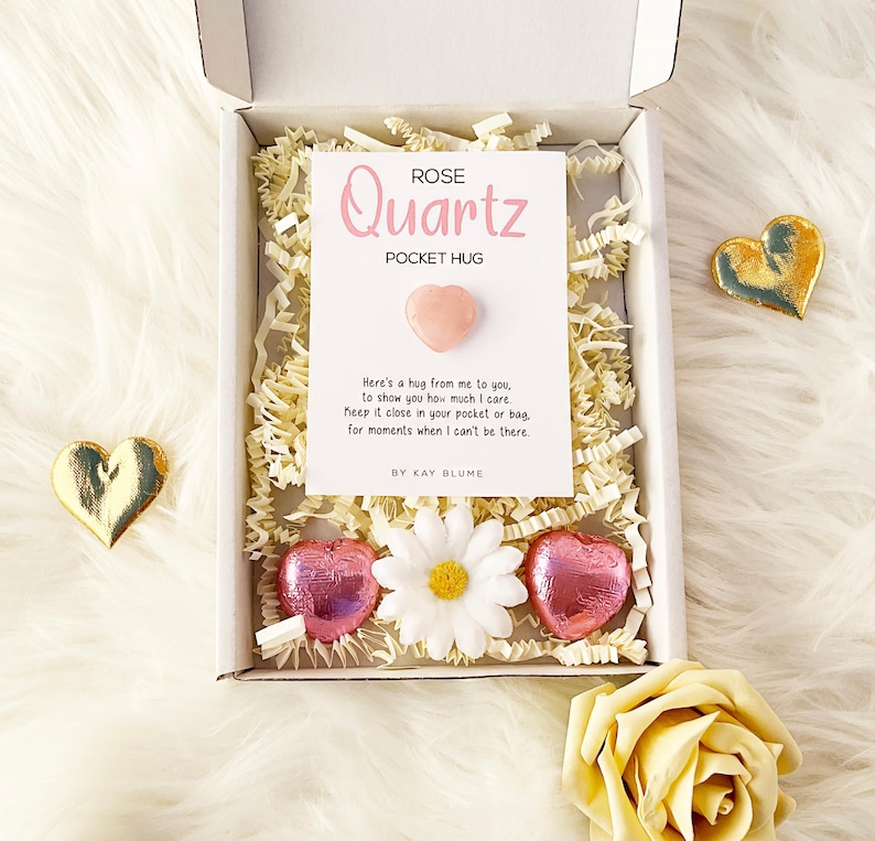 K&ouml;nnte beinhalten: Eine wei&szlig;e Geschenkbox mit einer wei&szlig;en Karte mit der Aufschrift Rose Quartz Pocket Hug und einem kleinen rosa herzf&ouml;rmigen Stein. Die Box enth&auml;lt geschreddertes Papier, ein wei&szlig;es G&auml;nsebl&uuml;mchen und zwei rosa folierte Herzschokoladen.