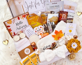 May include: A gift box labeled Just for You containing an autumn-themed card, honey peel-off mask, hand mask, lip balm, tea bags, hot chocolate, soap, white fuzzy socks, a white headband, a mustard yellow scrunchie, and various self-care items.