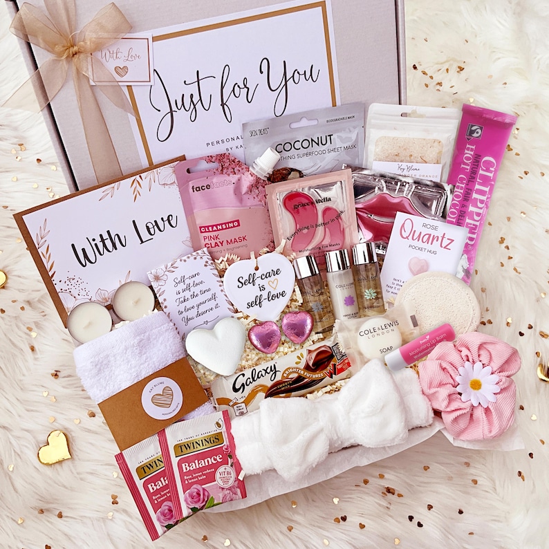 K&ouml;nnte beinhalten: Eine Geschenkbox mit der Aufschrift Just for You, die eine rosa Tonmaske, eine Kokosnuss-Tuchmaske, Augenmasken, Lippenbalsam, ein wei&szlig;es Stirnband, ein rosa Haargummi mit G&auml;nsebl&uuml;mchen, Twinings-Tee, einen Galaxy-Schokoriegel und Kerzen enth&auml;lt.