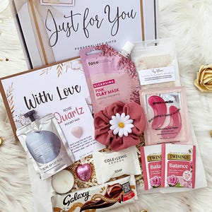 K&ouml;nnte beinhalten: Eine Geschenkbox mit einer Face Facts rosa Tonmaske, einem rosa Haargummi mit G&auml;nsebl&uuml;mchen, Grace & Stella Augenmasken, zwei Twinings Balance Teebeuteln, einem Galaxy Schokoriegel, einer Cole & Lewis Seife, einem Rosenquarz-Handschmeichler und 30 ml Shampoo.