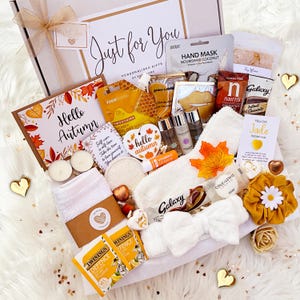 May include: A gift box with beauty and self-care products, including a 'Hello Autumn' card, a white bow, and a yellow scrunchie. The box contains a hand mask, chocolate, tea, and various skincare items.