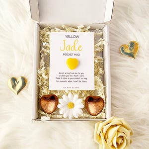 以下が含まれることがあります： YELLOW Jade POCKET HUGと書かれたカードと小さな黄色いハート型の石が入った白いギフトボックス。箱の中には紙の詰め物、2つの銅色のハート型のアイテム、白いデイジーのような花の装飾が入っています。