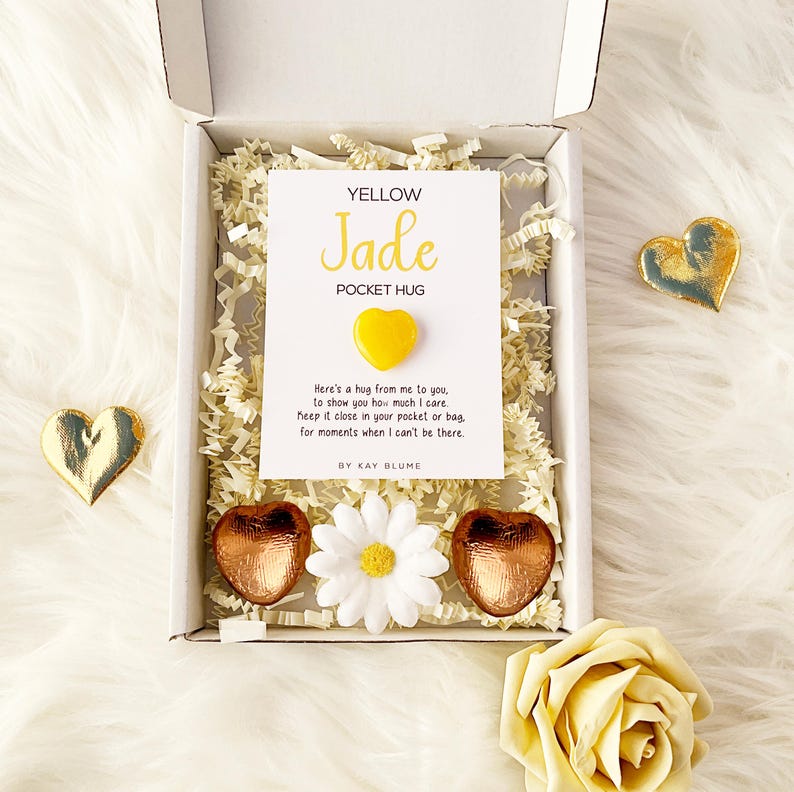 以下が含まれることがあります： YELLOW Jade POCKET HUGと書かれたカードと小さな黄色いハート型の石が入った白いギフトボックス。箱の中には紙の詰め物、2つの銅色のハート型のアイテム、白いデイジーのような花の装飾が入っています。