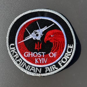 Op de afbeelding: Ronde patch met een zwarte achtergrond en witte stiksels. De patch toont een wit straalvliegtuig, een rode adelaar en de tekst "GHOST OF KYIV" en "UKRAINIAN AIR FORCE". Het ontwerp is geplaatst binnen een rode en witte cirkelvormige rand.