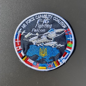 Può includere: Una toppa rotonda con la scritta "AIR FORCE CAPABILITY COALITION" e "F-16 Fighting Falcon". La toppa presenta un contorno blu dell'Ucraina e un jet. Il bordo è decorato con varie bandiere.
