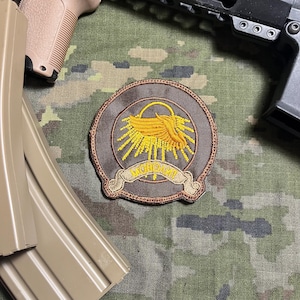 May include: A circular patch with a brown border and a central design featuring a yellow emblem resembling a stylized bird or wing, radiating sun rays. The patch includes a banner with text. The background is camouflage.