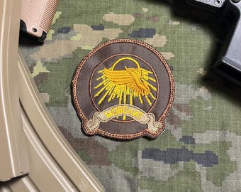 Op de afbeelding: Een ronde patch met een bruine rand en een centraal ontwerp met een geel embleem dat lijkt op een gestileerde vogel of vleugel, die zonnestralen uitstraalt. De patch bevat een banner met tekst. De achtergrond is camouflage.