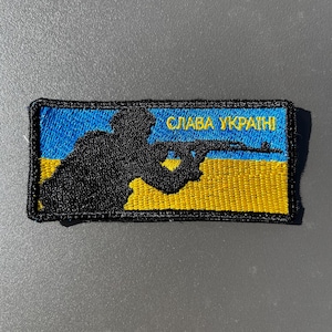以下が含まれることがあります： 兵士のシルエットがライフルを構えている刺繍パッチ。パッチは青と黄色の背景で、「СЛАВА УКРАЇНІ」の文字が黄色で書かれています。長方形のパッチには黒い縁取りがあります。