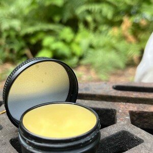May include: A small, round, black tin container with a lid. The container is filled with a yellow balm.