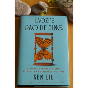 May include: A blue book cover with the title "Laozi's Dao De Jing" in black text. The cover features a butterfly inside an hourglass. The text below the butterfly reads "A new interpretation for a transformative time" and "Ken Liu".
