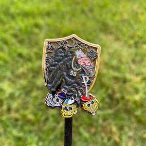 May include: A gold-colored pin featuring a black and white cartoon vulture wearing a chain with the number 98. The vulture is holding a sword and is surrounded by four football helmets in different colors.