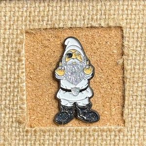 May include: A white glitter gnome with a black eye patch and a white beard is giving the middle finger with both hands. The gnome is wearing a white hat and a white shirt with a black belt. The gnome is standing on a brown corkboard.