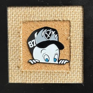 May include: A black and white enamel pin featuring a cartoon character wearing a black and white baseball cap with the number "87" on it. The character is peeking over a surface with a surprised expression.
