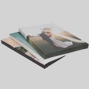 Op de afbeelding: Een stapel van drie rechthoekige canvasprints. Het bovenste canvas toont een foto van een koppel in trouwkleding dat elkaar omhelst in een veld. Het middelste canvas toont een blauwachtig landschap en het onderste canvas een zonsondergang.