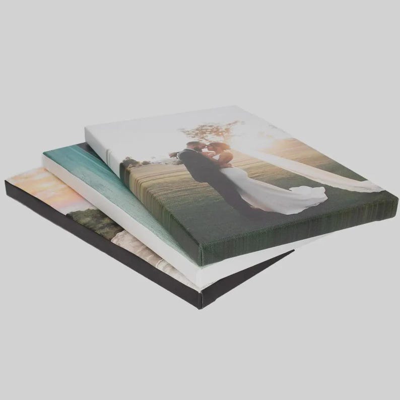 Op de afbeelding: Een stapel van drie rechthoekige canvasprints. Het bovenste canvas toont een foto van een koppel in trouwkleding dat elkaar omhelst in een veld. Het middelste canvas toont een blauwachtig landschap en het onderste canvas een zonsondergang.