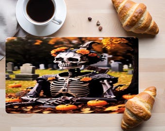 May include: A rectangular placemat featuring a printed illustration of a skeleton adorned with orange flowers, set in a graveyard with pumpkins and autumn leaves. The skeleton is positioned centrally, surrounded by tombstones and fall foliage.