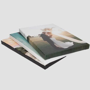 May include: Three rectangular canvas prints stacked on top of each other. The top canvas features a photograph of a couple in formal attire embracing in a field at sunset. The middle canvas shows a coastal landscape, and the bottom is black.