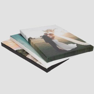 May include: Three rectangular canvas prints stacked on top of each other. The top canvas features a photograph of a couple in wedding attire embracing in a field. The middle canvas shows a landscape with blue tones, and the bottom canvas is black.