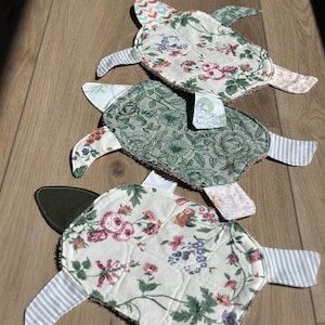 May include: Three fabric turtle-shaped items featuring quilted shells with floral patterns in shades of green, pink, and cream. Each turtle has patterned fabric flippers and heads in various prints including stripes, chevrons, and solid dark green.