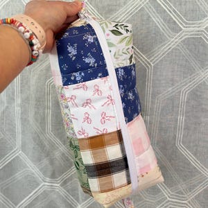 May include: A patchwork fabric bag with a variety of floral and patterned fabrics in shades of blue, pink, green, and brown. The bag is held by a loop and secured with a white elastic band.