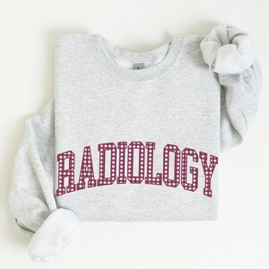May include: A light gray crewneck sweatshirt featuring the word RADIOLOGY printed in a maroon and white gingham check pattern across the chest. The garment has long sleeves with ribbed cuffs and a classic round neckline in a soft fleece fabric.