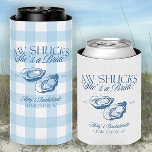 May include: Two white and blue can coolers with a gingham pattern. The coolers feature a blue and white illustration of oysters and the text "Aw Shucks She's a Bride!" with the text "Abby's Bachelorette Charleston, SC" and the years "EST. 2025".