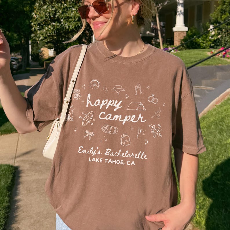 May include: A brown short-sleeved t-shirt featuring white line art graphics of camping gear and the text "happy camper", "Emily's Bachelorette", and "LAKE TAHOE, CA". The shirt has a crew neckline and a loose fit, worn with a white shoulder bag.