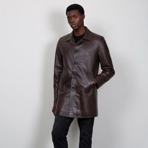 May include: A brown leather jacket with a button-up front and long sleeves. The jacket is worn by a person with dark skin and black hair. The person is wearing black pants and dark green boots.