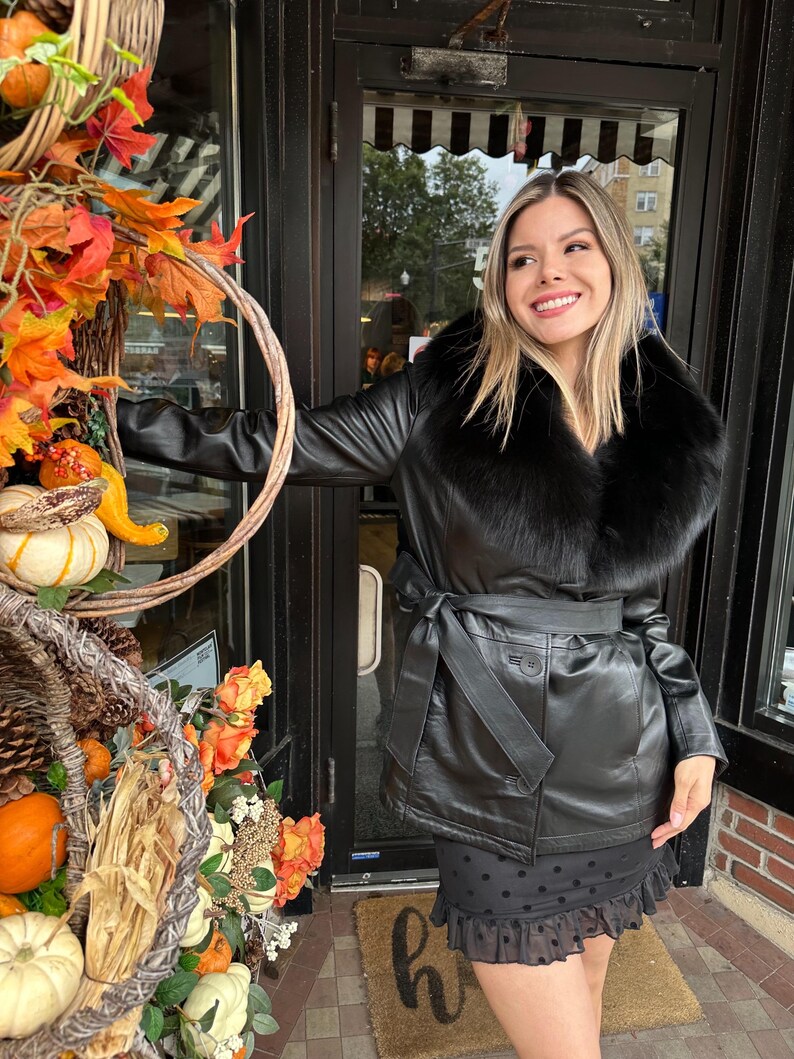 May include: A black leather jacket with a large black fur collar and a black belt. The jacket is belted at the waist and has a button closure. The model is wearing a black polka dot skirt.