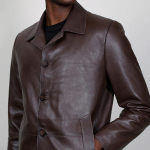 May include: A brown leather jacket with a button-up front and a long, straight cut. The jacket has a collar and two pockets.