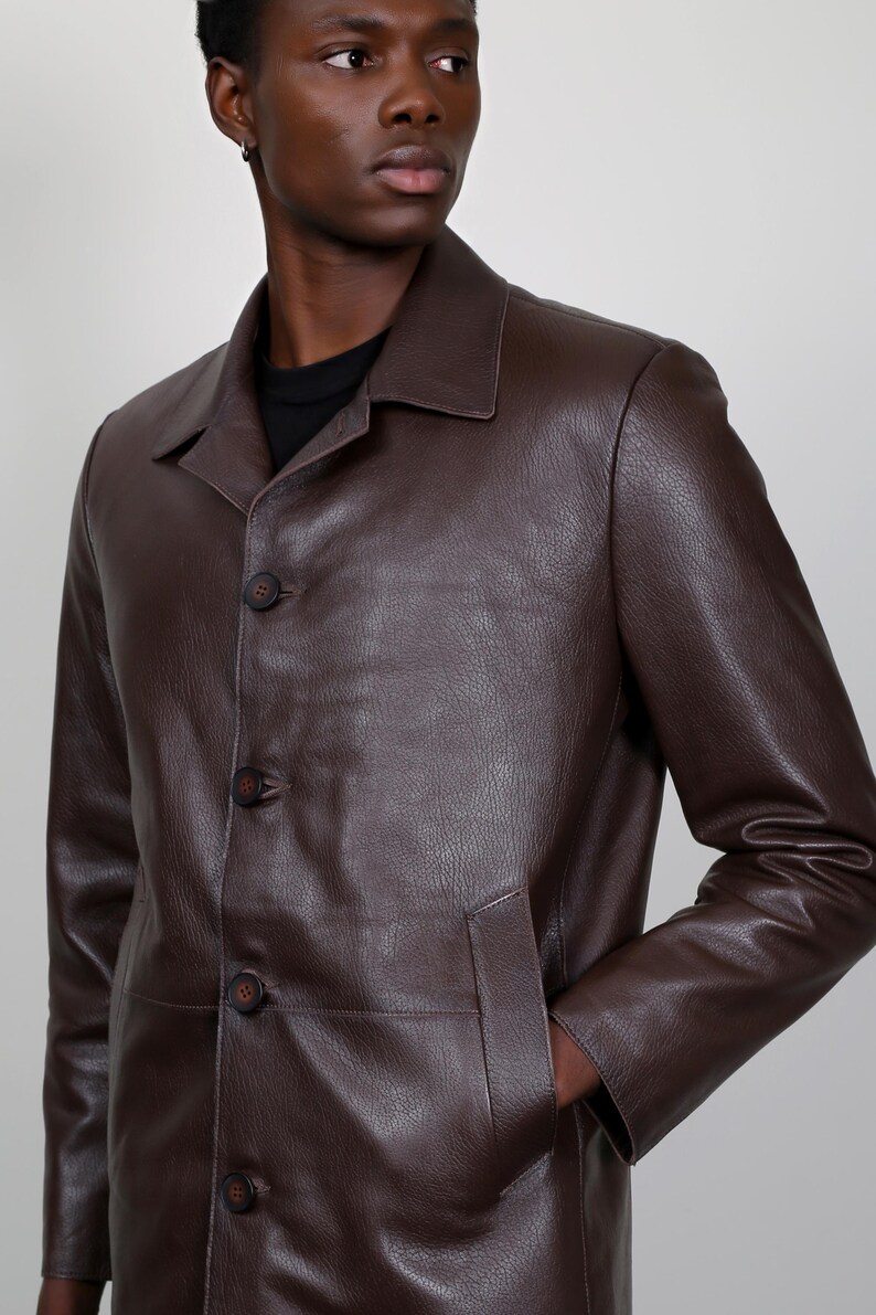 May include: A brown leather jacket with a button-up front and a long, straight cut. The jacket has a collar and two pockets.