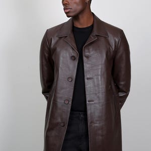 May include: A brown leather jacket with a collared neckline and button closure. The jacket has a long, fitted silhouette and is worn by a person with dark skin.