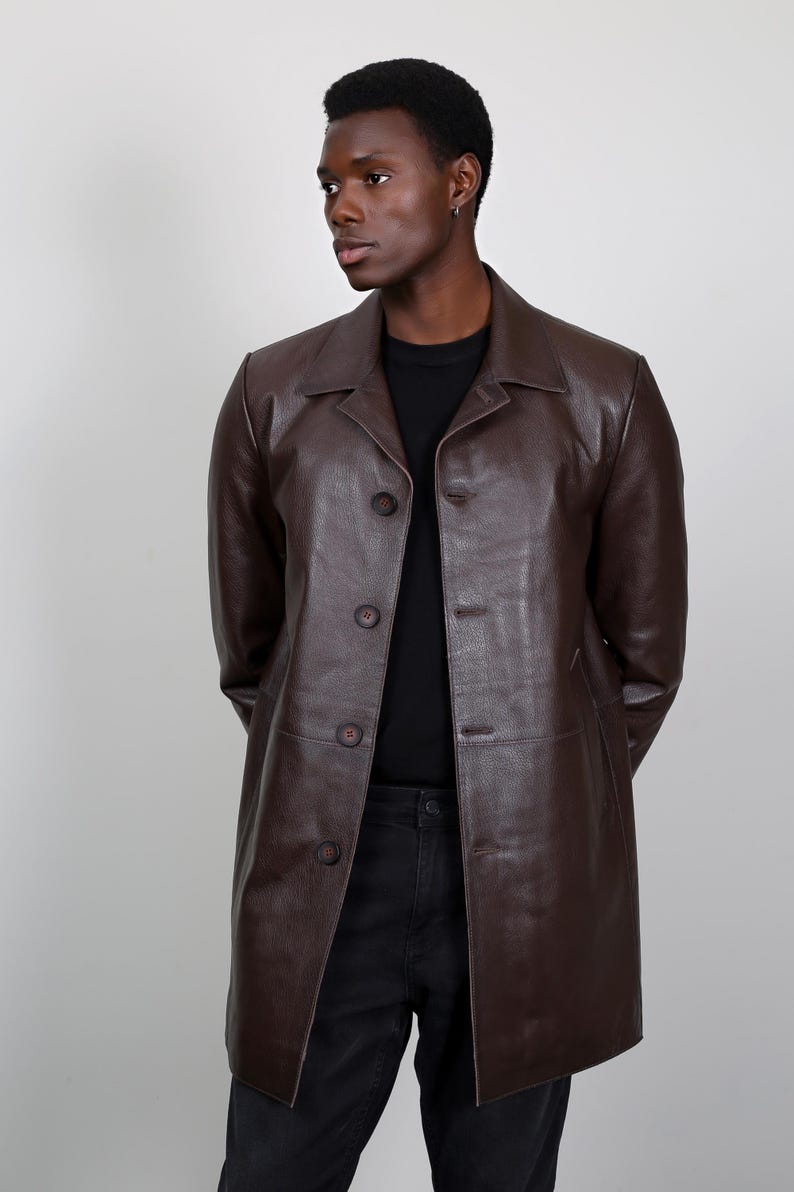 May include: A brown leather jacket with a collared neckline and button closure. The jacket has a long, fitted silhouette and is worn by a person with dark skin.