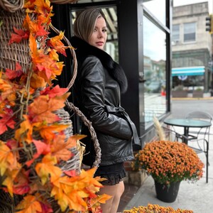 May include: A woman wearing a black leather jacket with a fur collar stands in front of a storefront. The jacket has a belt and a fur trim around the collar. The woman is standing in front of a storefront with a sign that says "Sweet Kitchen".