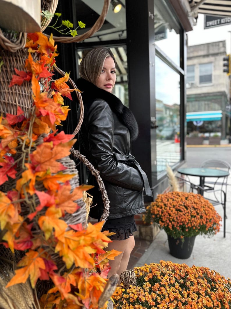 May include: A woman wearing a black leather jacket with a fur collar stands in front of a storefront. The jacket has a belt and a fur trim around the collar. The woman is standing in front of a storefront with a sign that says "Sweet Kitchen".
