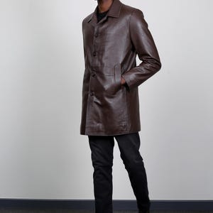 May include: A brown leather coat with a button closure and a collar. The coat is worn by a person with dark skin and black hair. The person is wearing black pants and dark green boots.