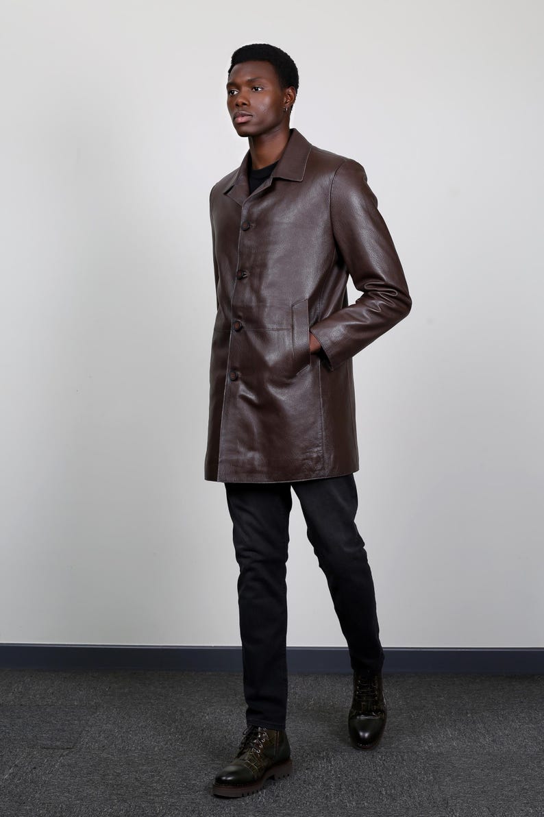 May include: A brown leather coat with a button closure and a collar. The coat is worn by a person with dark skin and black hair. The person is wearing black pants and dark green boots.
