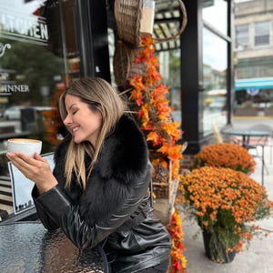 May include: A woman wearing a black leather jacket with a fur collar sits at a table outside a cafe, holding a cup of coffee. The cafe has a sign that reads "Kitchen" and "Dinner".