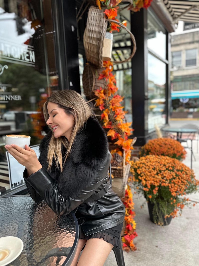 May include: A woman wearing a black leather jacket with a fur collar sits at a table outside a cafe, holding a cup of coffee. The cafe has a sign that reads "Kitchen" and "Dinner".