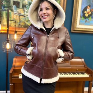 Peut inclure: Une veste en cuir marron avec une doublure en peau de mouton blanche et une capuche. La veste a une fermeture &eacute;clair et deux poches.