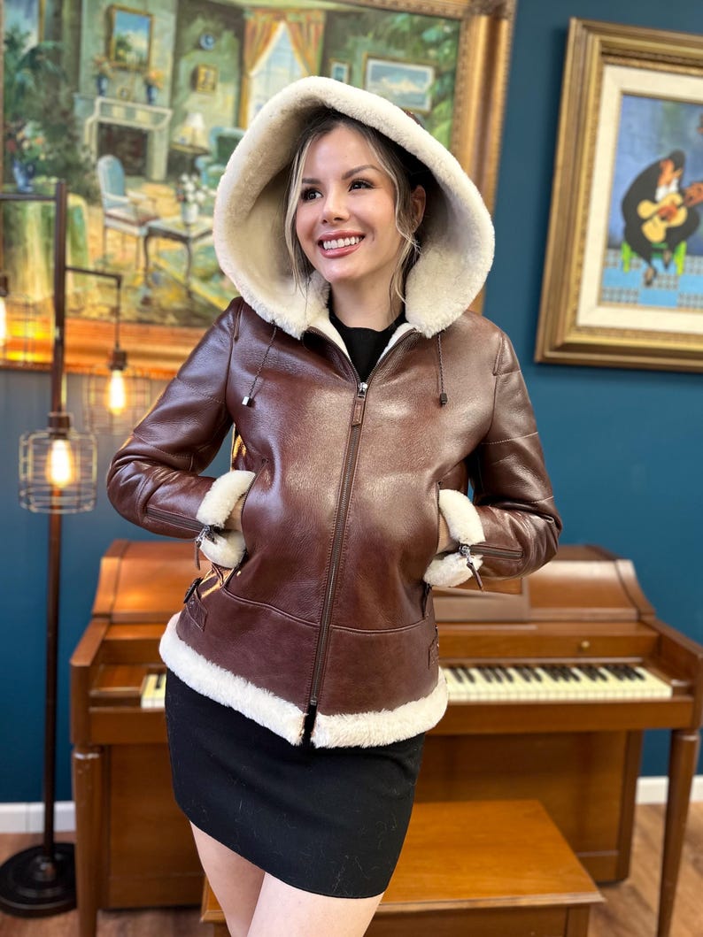 Peut inclure: Une veste en cuir marron avec une doublure en peau de mouton blanche et une capuche. La veste a une fermeture &eacute;clair et deux poches.